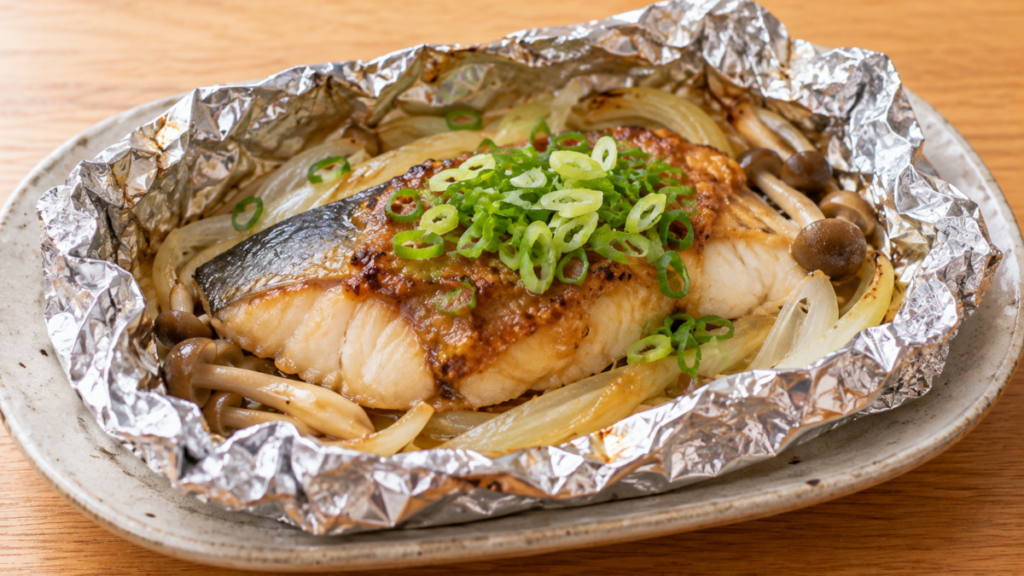 ブリの切り身を玉ねぎやきのこと一緒にホイルで包み、ネギ味噌だれと青ねぎをのせて焼いた料理写真